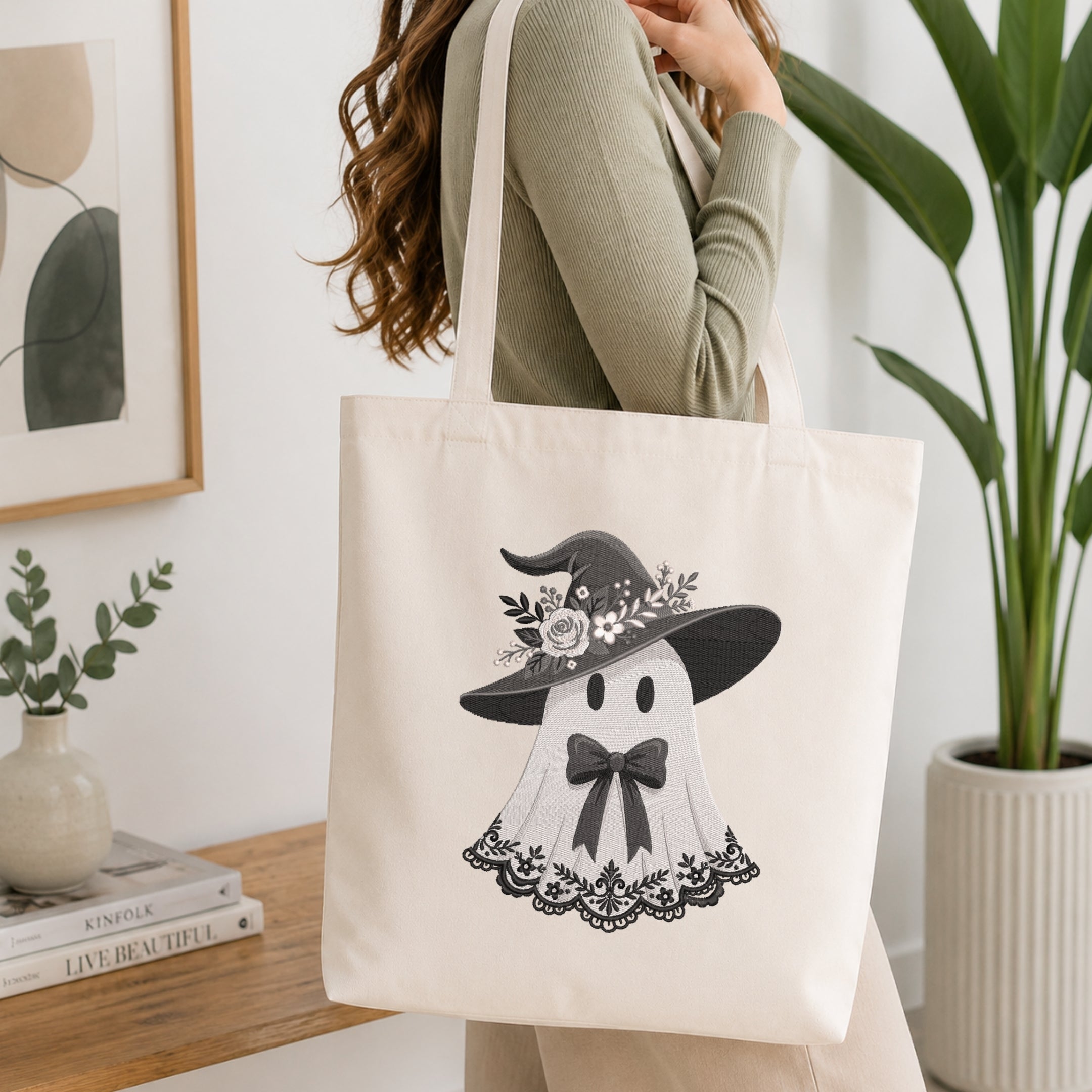 A woman wearing a sage green ribbed sweater carries a cream canvas tote bag with a black and white graphic of a ghost wearing a witch hat. The scene is set in a bright, modern room with a wooden table, books, a small plant, and a large potted plant in the background.