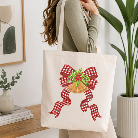 A woman wearing a sage green ribbed sweater carries a cream canvas tote bag with a red and white gingham bow, gold bells, and green holly design on the front, set against a bright, modern indoor background with a potted plant and framed art.
