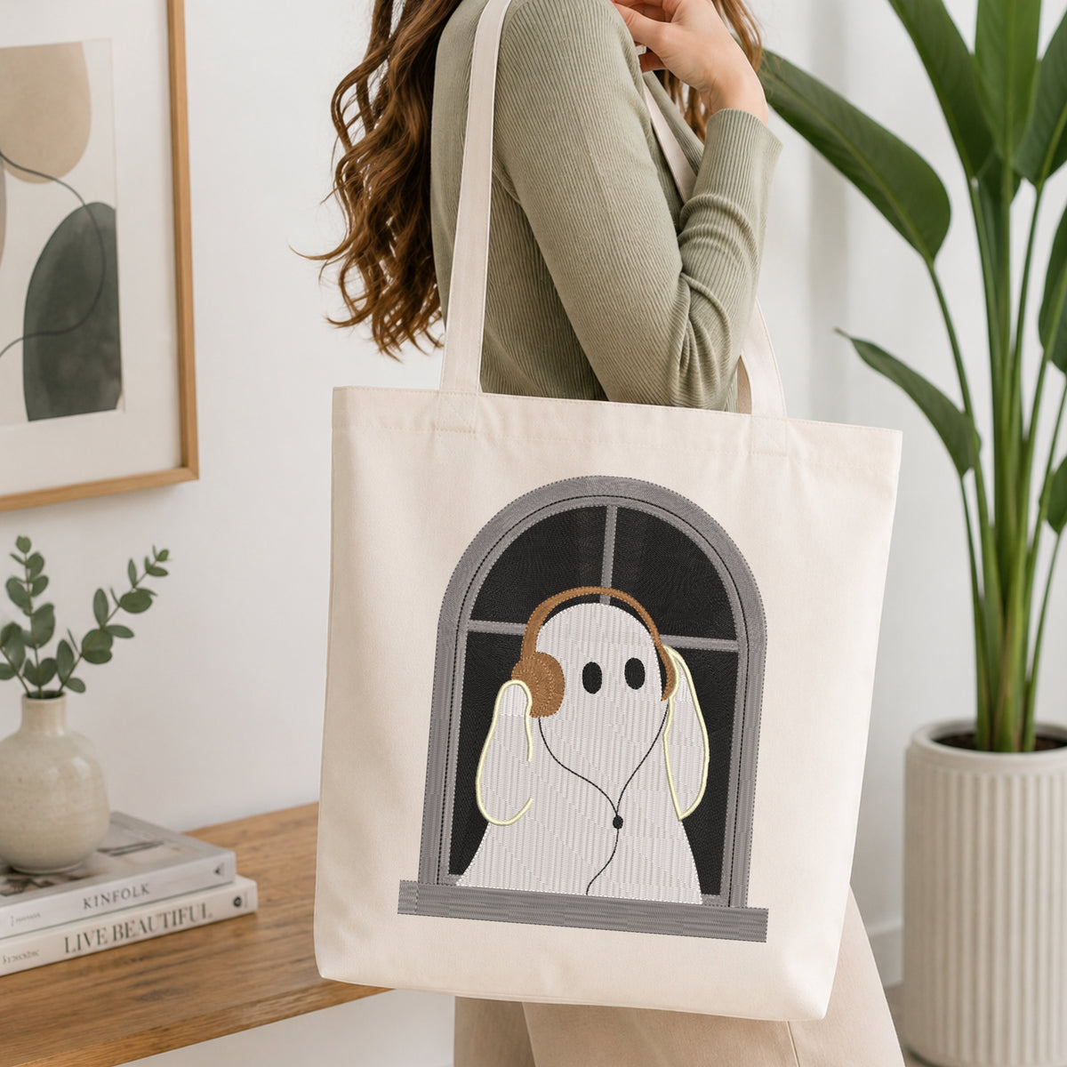 A woman wearing a sage green ribbed sweater carries a cream canvas tote bag with an embroidered design of a white ghost wearing headphones, seen in a bright, modern room with a wooden table and potted plants.