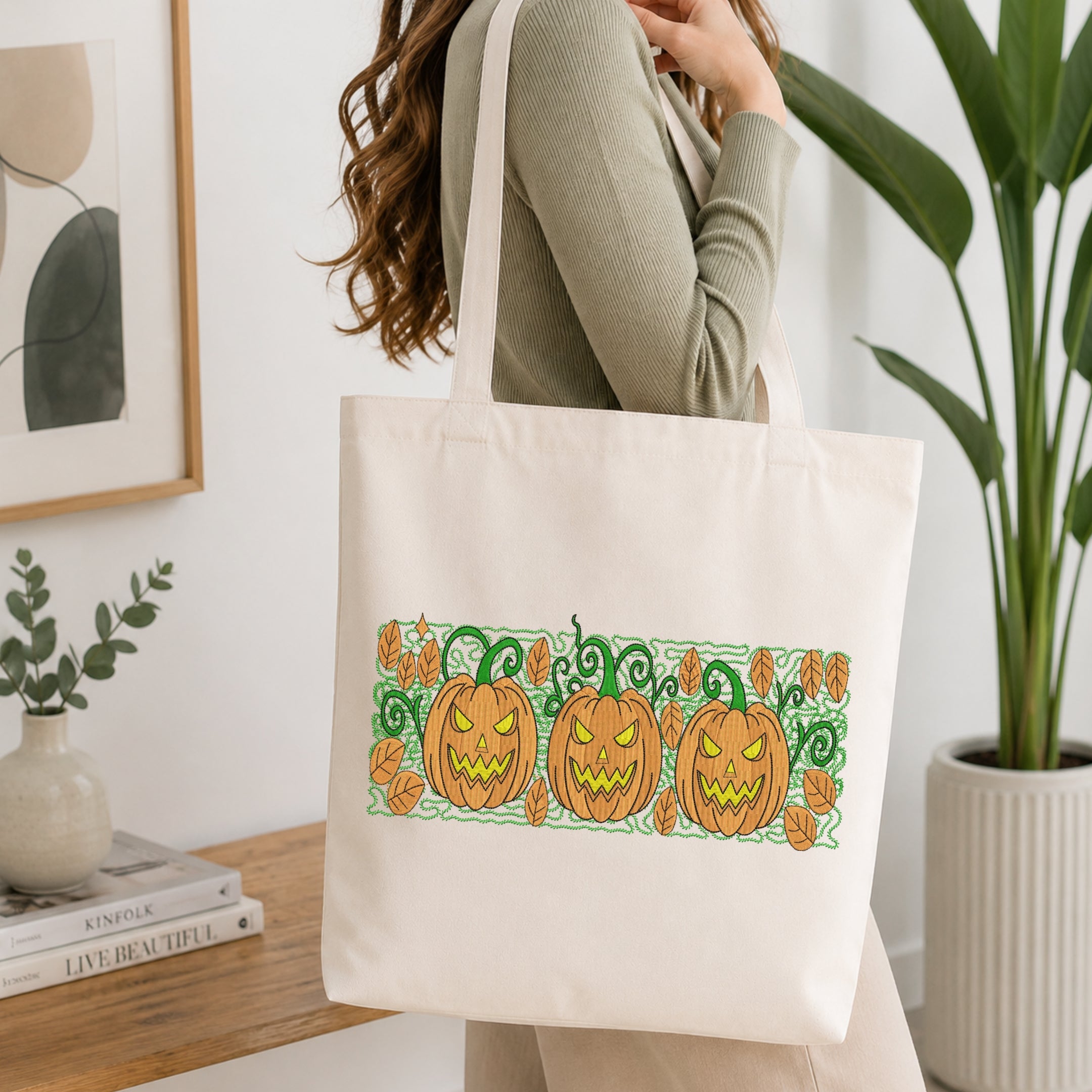 A woman wearing a sage green ribbed sweater carries a cream canvas tote bag with a horizontal graphic of three orange jack-o'-lantern pumpkins with green stems and leaves on a white background.