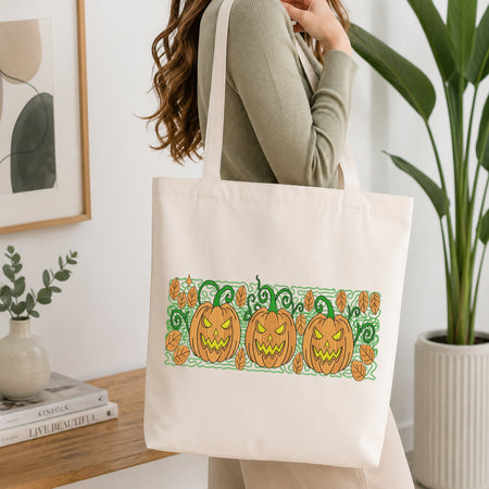 A woman wearing a sage green ribbed sweater carries a cream canvas tote bag with a horizontal graphic of three orange jack-o'-lantern pumpkins with green stems and leaves on a white background.
