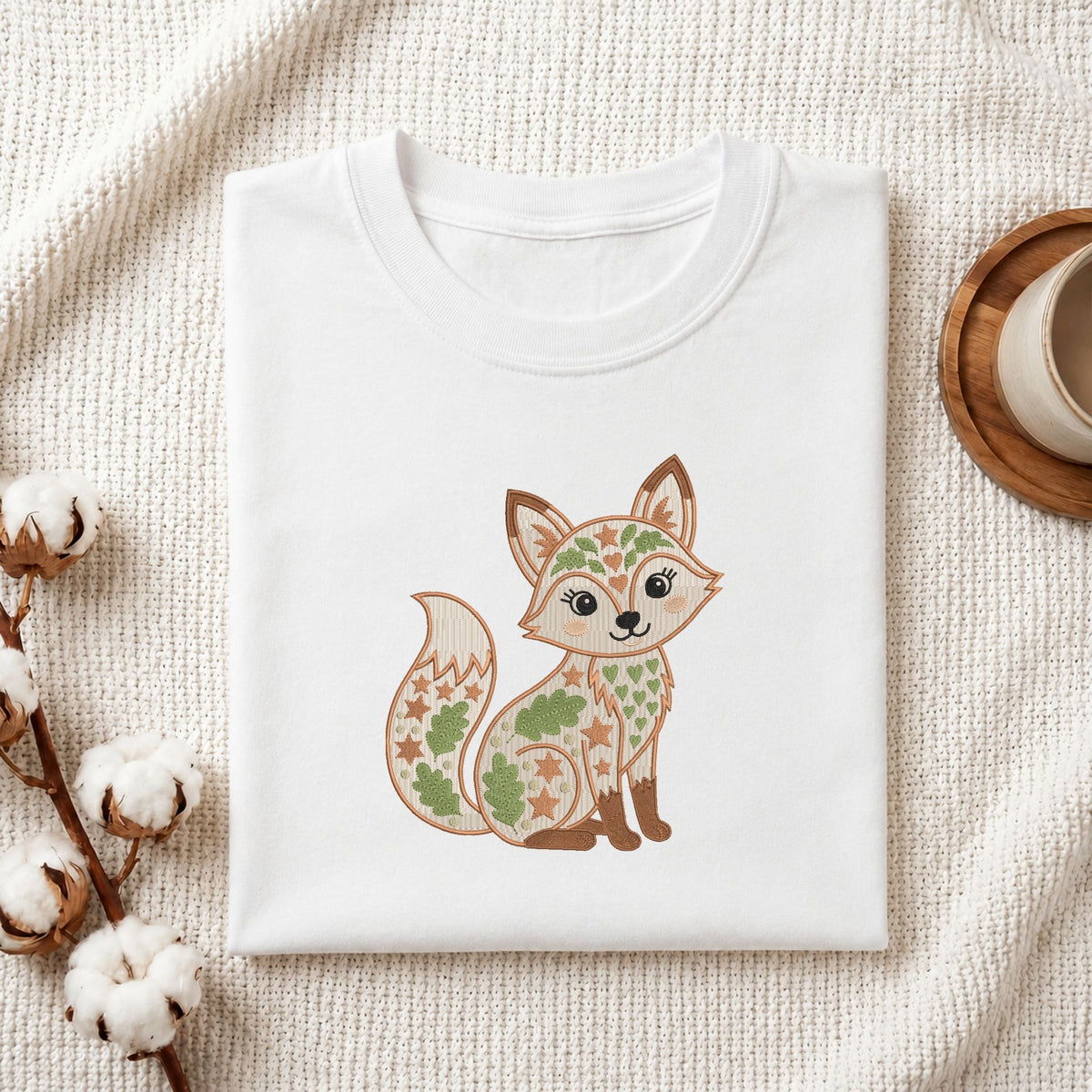 Folded white cotton t-shirt mockup featuring a cute folk art fox machine embroidery design, styled with cotton stems and a wooden cup.   