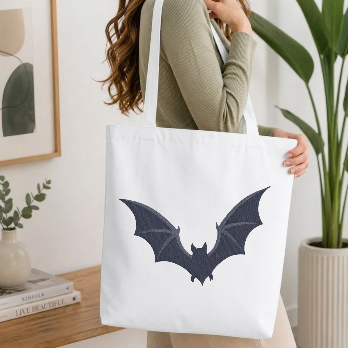 A woman wearing a sage green ribbed sweater carries a white canvas tote bag with a dark navy bat graphic on the front, set in a bright, modern room with a wooden table and potted plants.