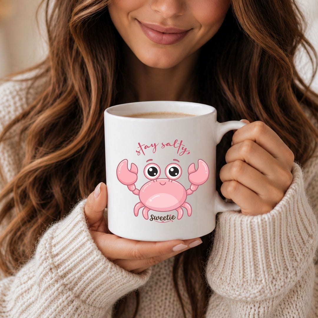 A woman with long brown hair holding a white ceramic mug featuring a cartoon pink crab graphic and the text "stay salty, Sweetie".