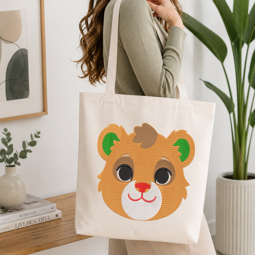 A person wearing a sage green ribbed sweater carries a cream canvas tote bag featuring a cute embroidered-style brown bear face with green ears and a pink nose, set against a bright, modern room with a wooden table and potted plants.