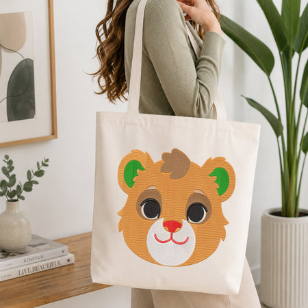 A person wearing a sage green ribbed sweater carries a cream canvas tote bag featuring a cute embroidered-style brown bear face with green ears and a pink nose, set against a bright, modern room with a wooden table and potted plants.