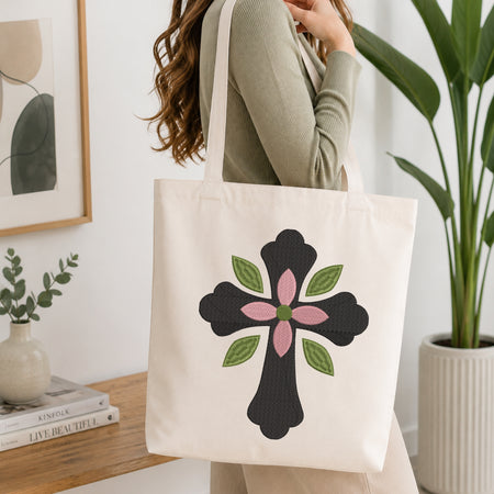 A woman wearing a sage green ribbed long-sleeve top carries a cream canvas tote bag with a black cross and pink flower design printed on the front, set in a bright, modern room with a wooden table and potted plants.