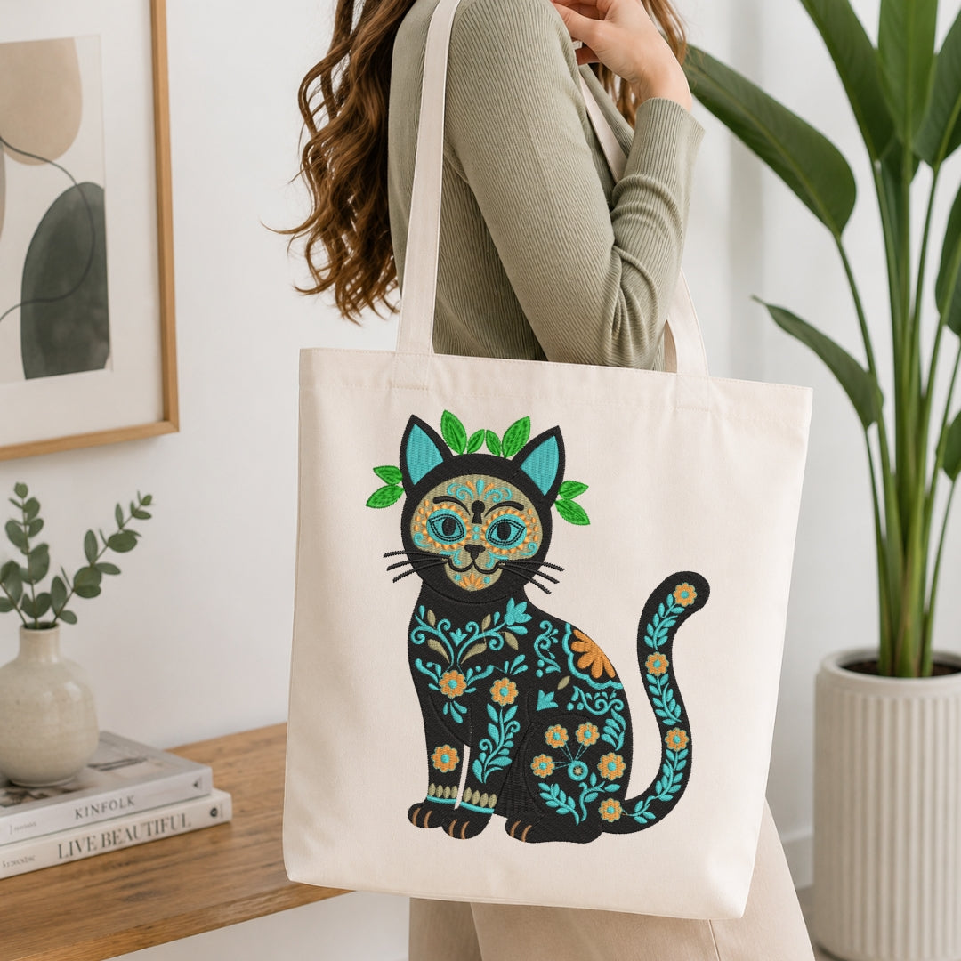 A person wearing a sage green ribbed sweater carries a cream canvas tote bag decorated with a colorful Dia de los Muertos-style cat design. The background shows a bright, modern room with a wooden table, books, a framed print, and a potted plant.