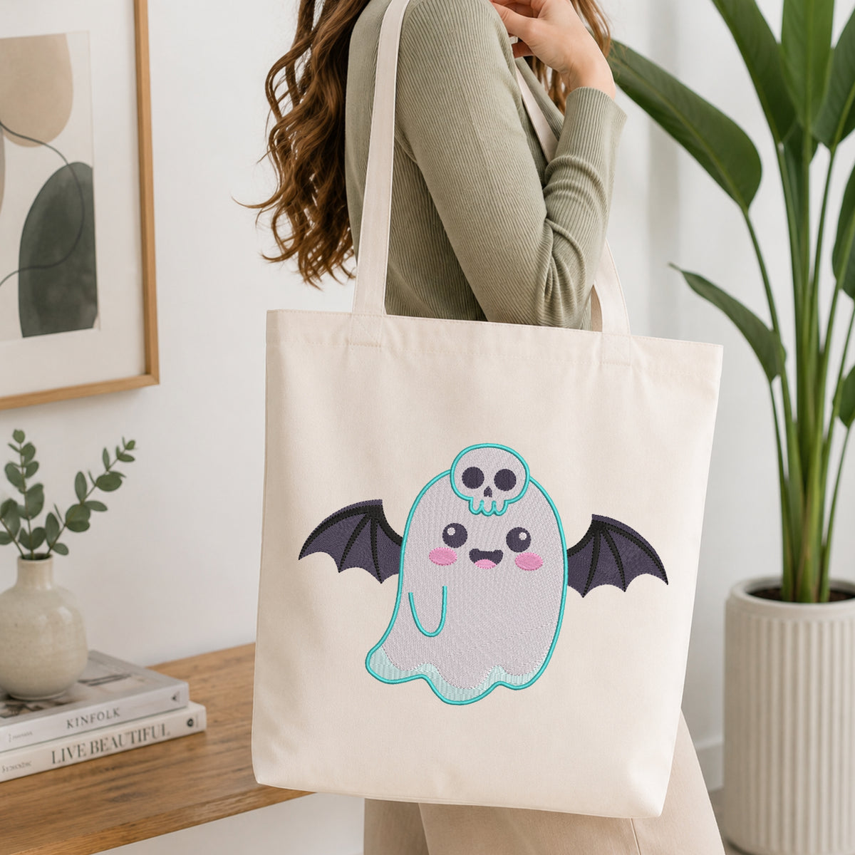 An off-white canvas tote bag with a cute embroidered bat ghost design, carried by a woman in a home setting with a wooden table and potted plants in the background.