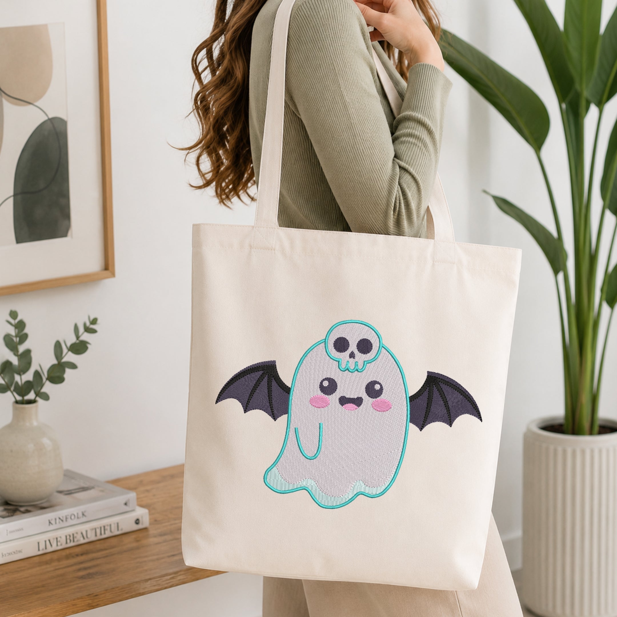 An off-white canvas tote bag with a cute embroidered bat ghost design, carried by a woman in a home setting with a wooden table and potted plants in the background.