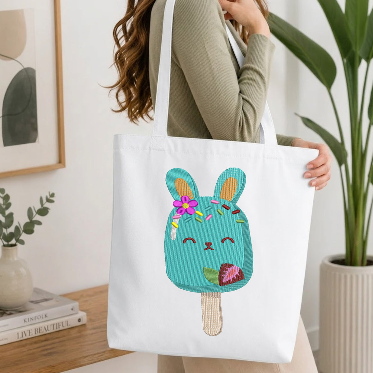 A white canvas tote bag with a cute embroidered turquoise bunny popsicle design, carried by a woman in a ribbed sage green top in a bright, modern room.