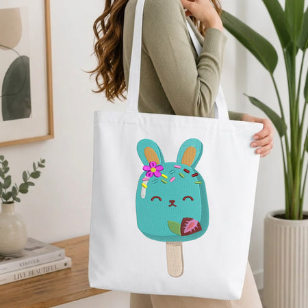 A white canvas tote bag with a cute embroidered turquoise bunny popsicle design, carried by a woman in a ribbed sage green top in a bright, modern room.