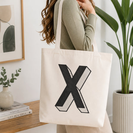 A woman wearing a sage green ribbed long-sleeve top carries a cream canvas tote bag with a large black 3D-effect letter 'X' printed on the front, set in a bright, modern room with a wooden table, framed art, and a potted plant.
