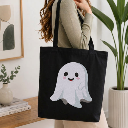 Woman carrying a black canvas tote bag featuring a cute smiling ghost machine embroidery design in a bright, modern interior setting