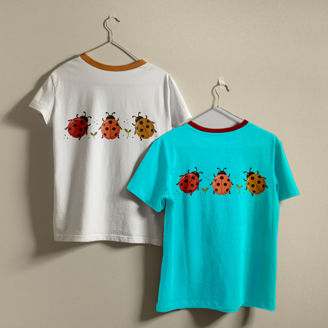 Two children's short-sleeve ringer T-shirts hanging on hangers against a plain beige wall. The back of one white shirt has a tan collar trim and a row of ladybug illustrations across the back.