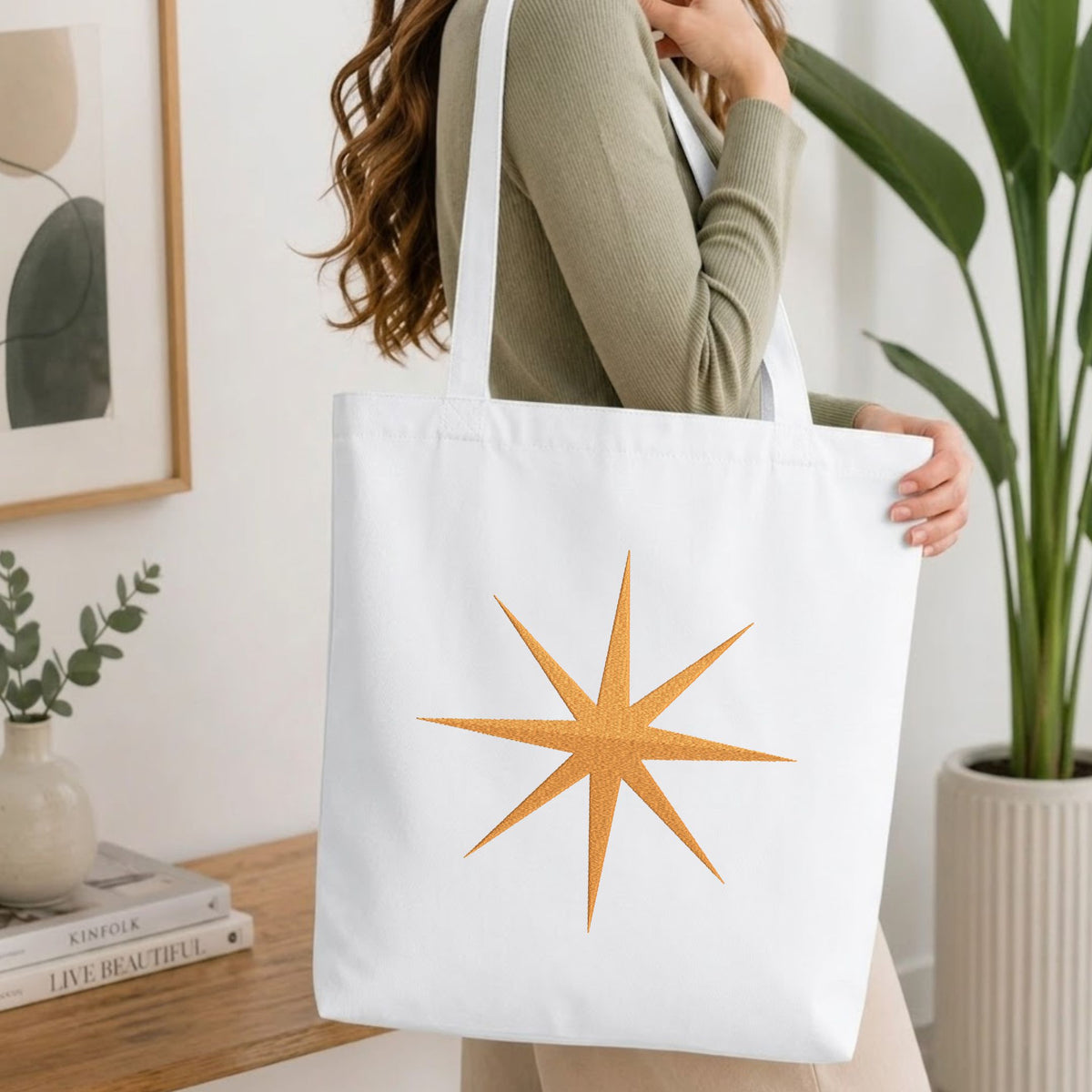 Folded white cotton t-shirt mockup featuring a golden starburst machine embroidery design, styled with cotton stems and a wooden cup