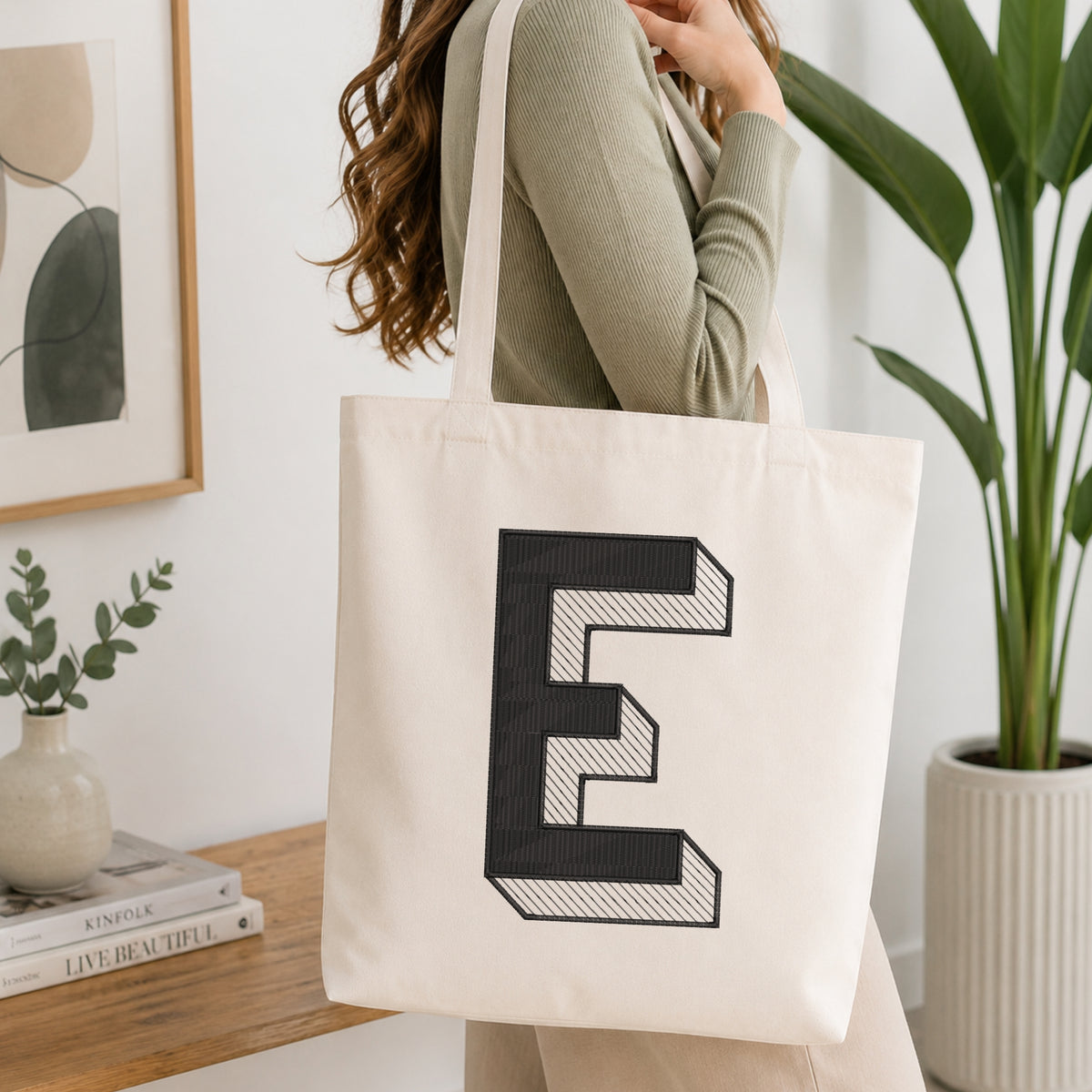 A woman wearing a sage green ribbed top carries a cream canvas tote bag with a large black embroidered letter 'E' on the front, set in a bright, modern room with a wooden table, framed art, and a potted plant.