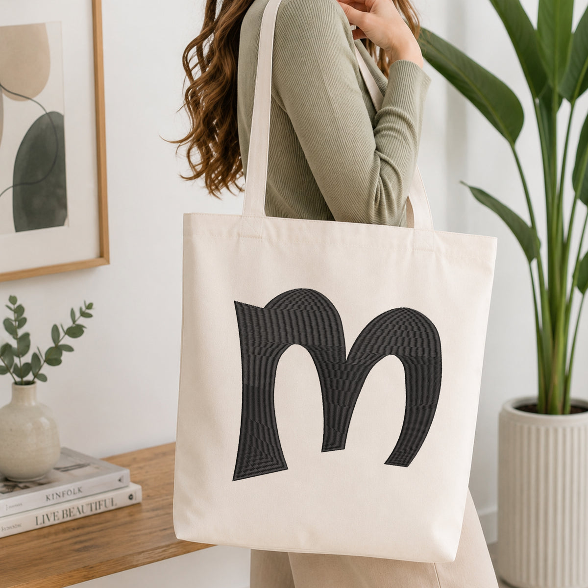 A woman wearing a sage green ribbed long-sleeve top carries a cream canvas tote bag with a large black textured letter 'm' printed on the front, set in a bright, modern room with a wooden console table, framed art, and a potted plant.