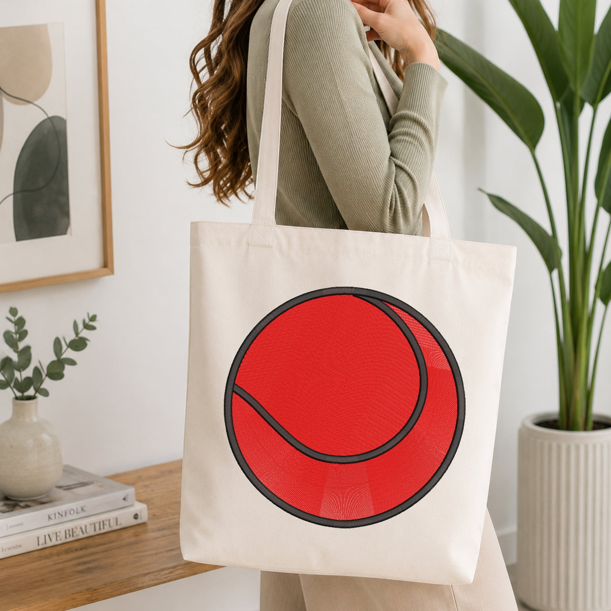 A woman wearing a sage green ribbed sweater carries a cream canvas tote bag with a large red tennis ball graphic outlined in black. The scene is set in a bright, modern room with a wooden table, books, a framed print, and a potted plant.