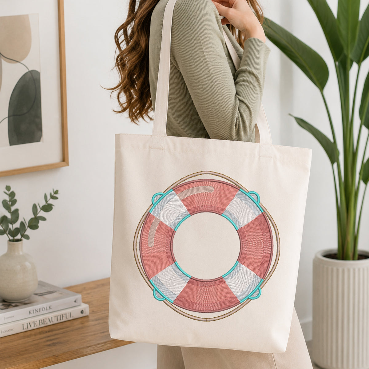 A woman wearing a sage green ribbed sweater carries a cream canvas tote bag with a pink, white, and teal life preserver graphic printed on the front, set in a bright, modern room with a wooden table and potted plants.