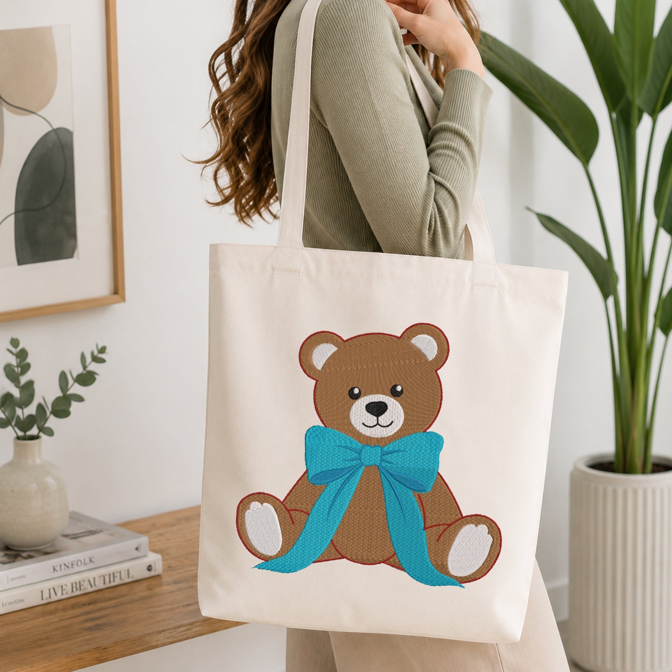 A person holding a natural canvas tote bag printed with an embroidered brown teddy bear design wearing a bright blue bow, set against an interior background with green plants.