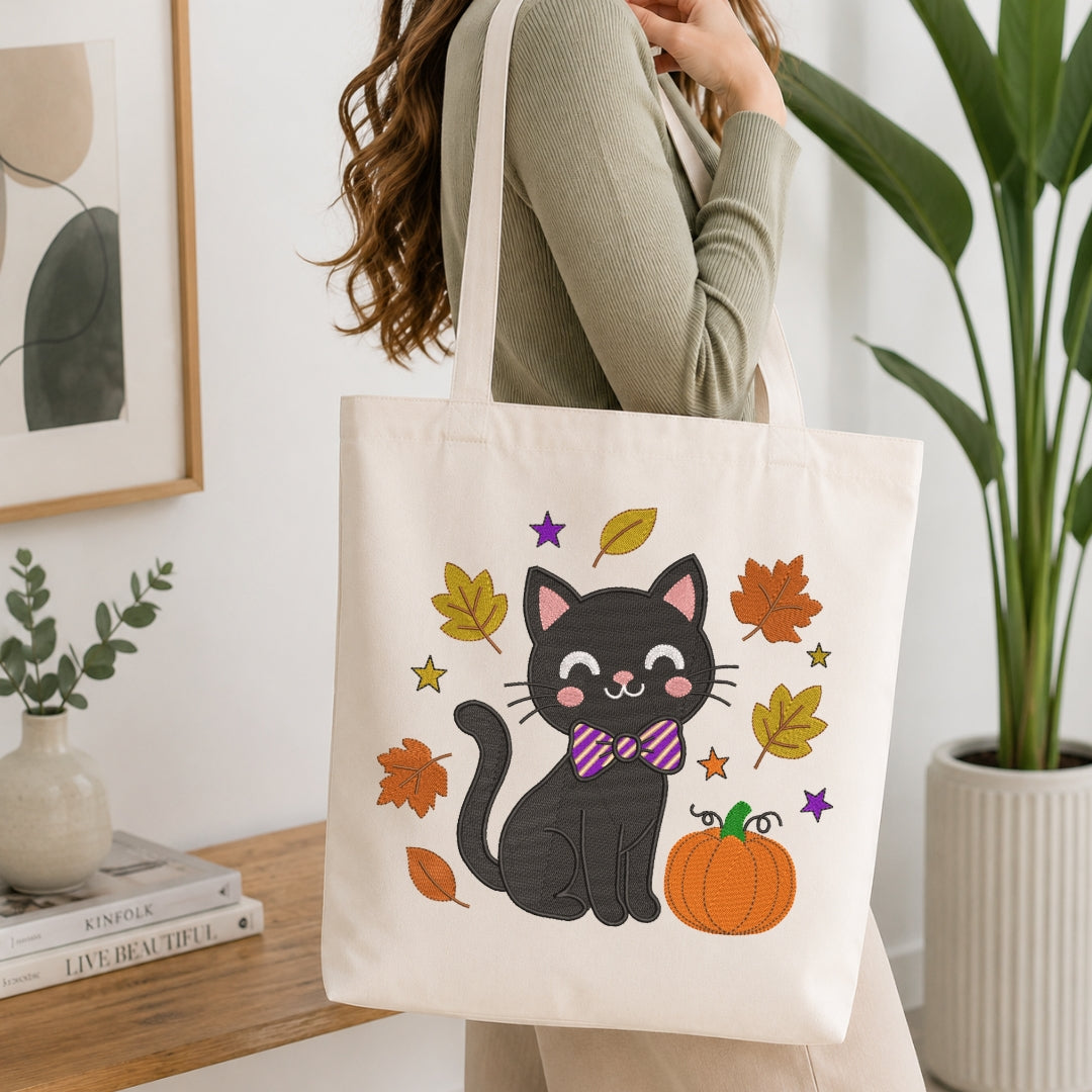 A woman wearing a sage green ribbed sweater carries a cream canvas tote bag. The bag features a cute cartoon illustration of a black cat wearing a purple and pink striped bow tie, surrounded by orange and yellow autumn leaves, stars, and a pumpkin.