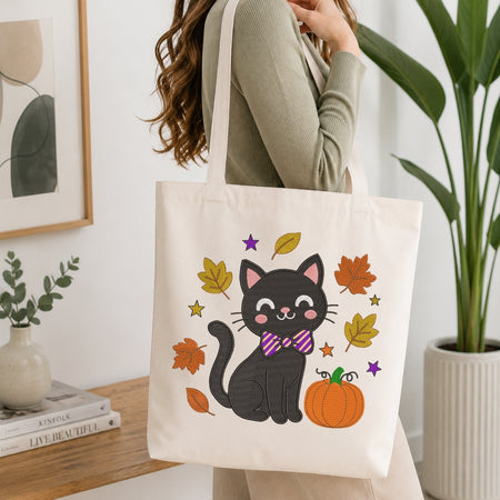 A woman wearing a sage green ribbed sweater carries a cream canvas tote bag. The bag features a cute cartoon illustration of a black cat wearing a purple and pink striped bow tie, surrounded by orange and yellow autumn leaves, stars, and a pumpkin.