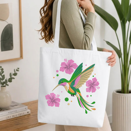 A woman holds a white canvas tote bag featuring a colorful illustration of a hummingbird and pink hibiscus flowers, set in a bright, modern room with a potted plant and wooden table.