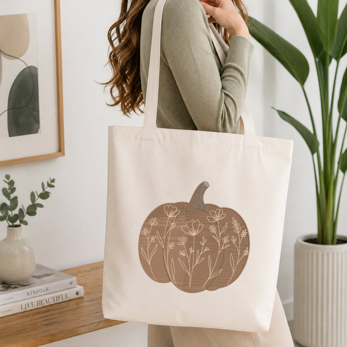 A woman wearing a sage green ribbed sweater carries a cream canvas tote bag with a brown pumpkin graphic featuring white floral line art, set in a bright, modern room with a wooden table and potted plants.