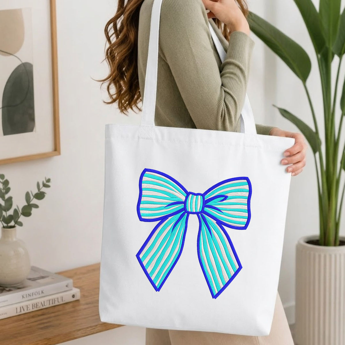 Woman carrying a white natural canvas tote bag featuring an embroidered blue and white striped bow design in a bright, modern interior setting.