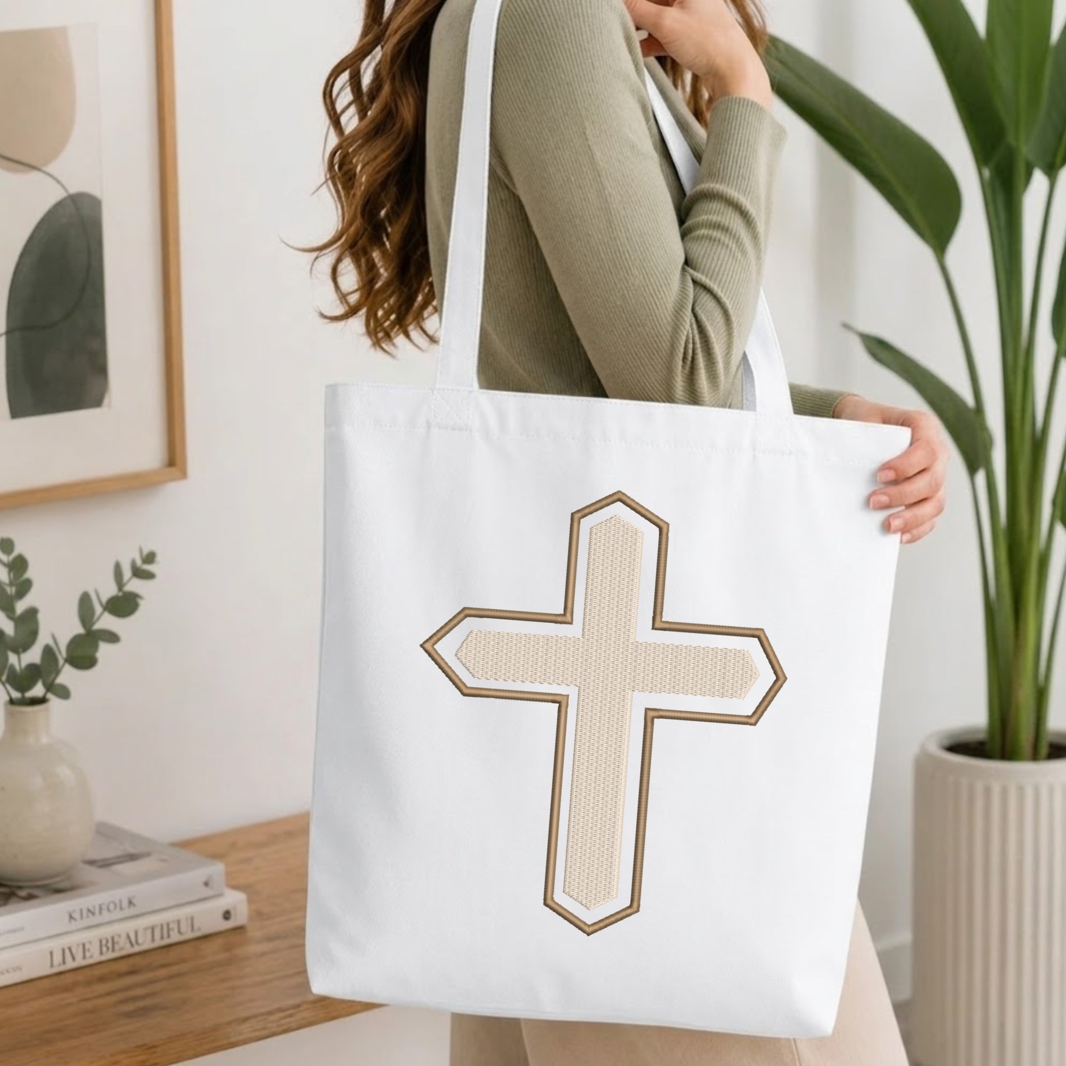 A person wearing a green top holds up a white canvas tote bag printed with the textured beige and brown Christian cross embroidery design, set against a bright, minimalist room background with indoor plants.