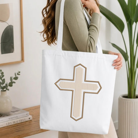 A person wearing a green top holds up a white canvas tote bag printed with the textured beige and brown Christian cross embroidery design, set against a bright, minimalist room background with indoor plants.