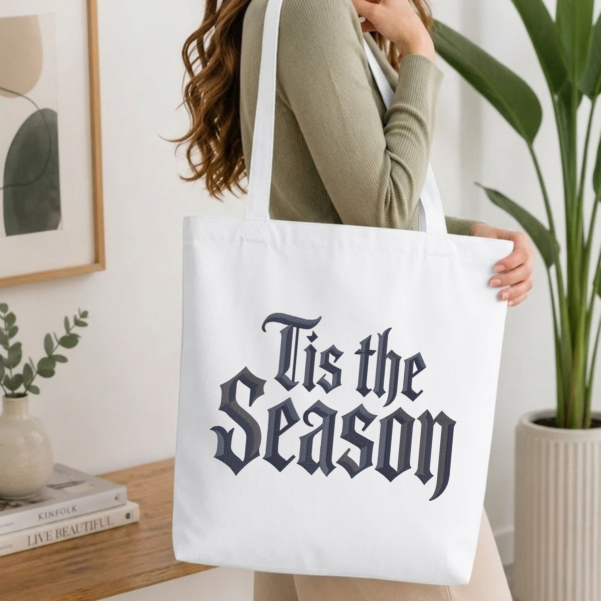 A woman wearing a sage green ribbed long-sleeve top carries a white canvas tote bag with the phrase 'Tis the Season' printed in a dark navy gothic font. The photo is taken in a bright, modern room with a wooden table, books, a framed print, and a potted plant.