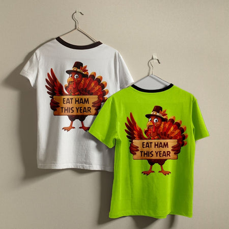 Two short-sleeve ringer T-shirts hanging on hangers against a plain beige wall. The left shirt is white with a black collar trim and shows the back graphic of a cartoon turkey holding a sign that reads 'EAT HAM THIS YEAR'.