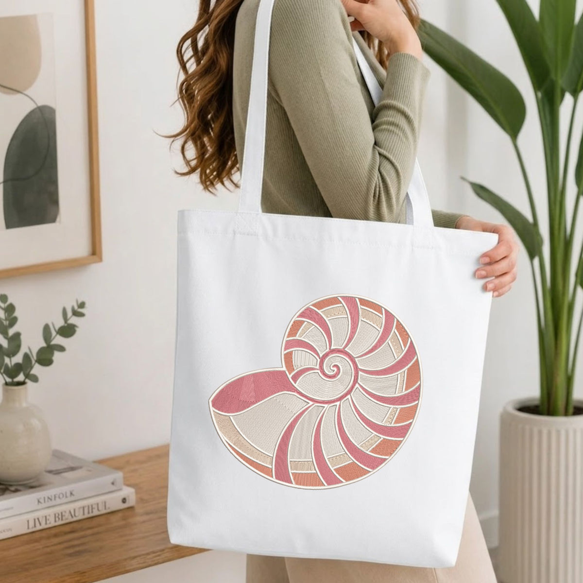A woman wearing a sage green ribbed sweater carries a white canvas tote bag with a large pink and beige nautilus shell graphic on the front, set in a bright, modern room with a wooden table and potted plants.