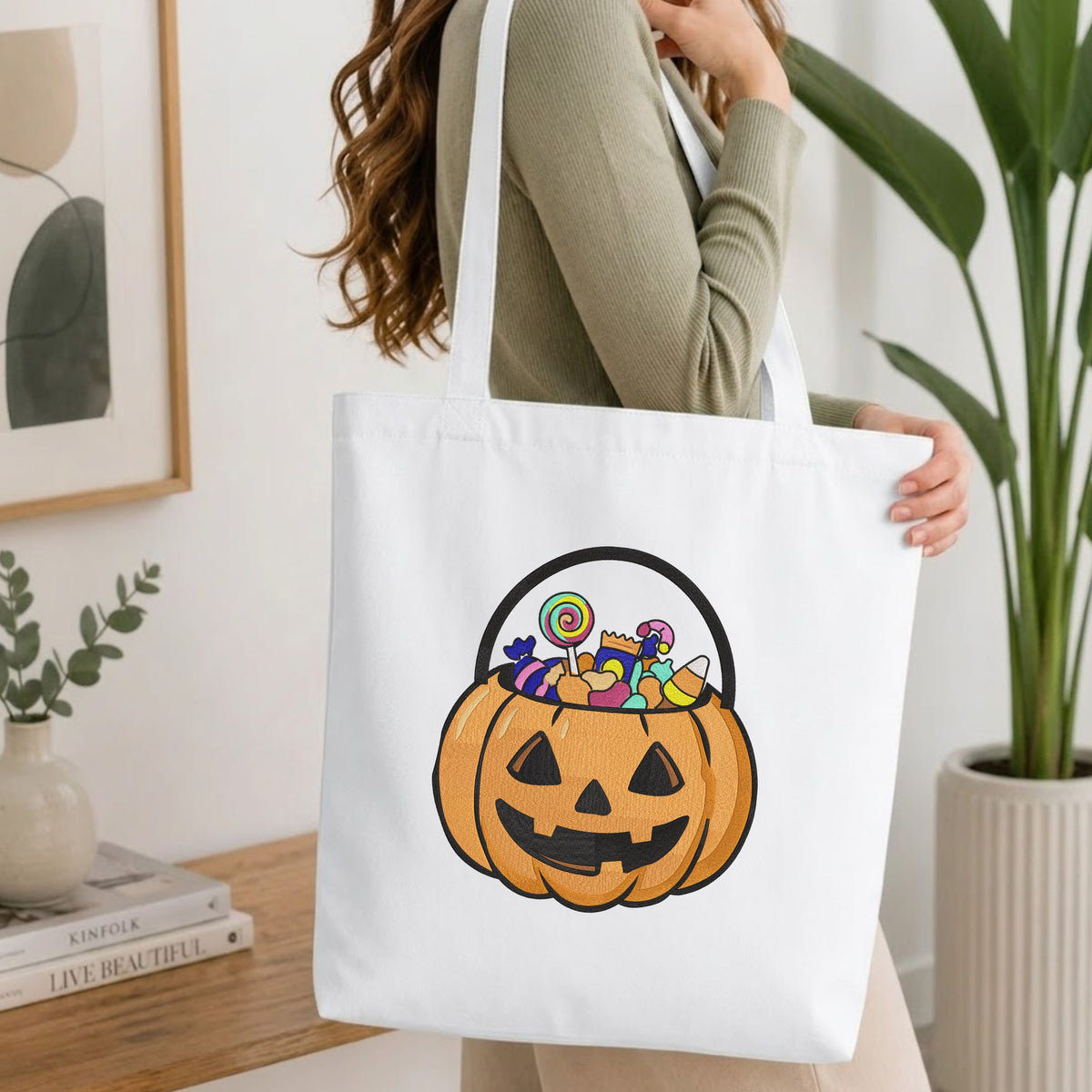  Woman carrying a white natural canvas tote bag featuring a jack-o'-lantern candy bucket machine embroidery design in a bright, modern interior setting   