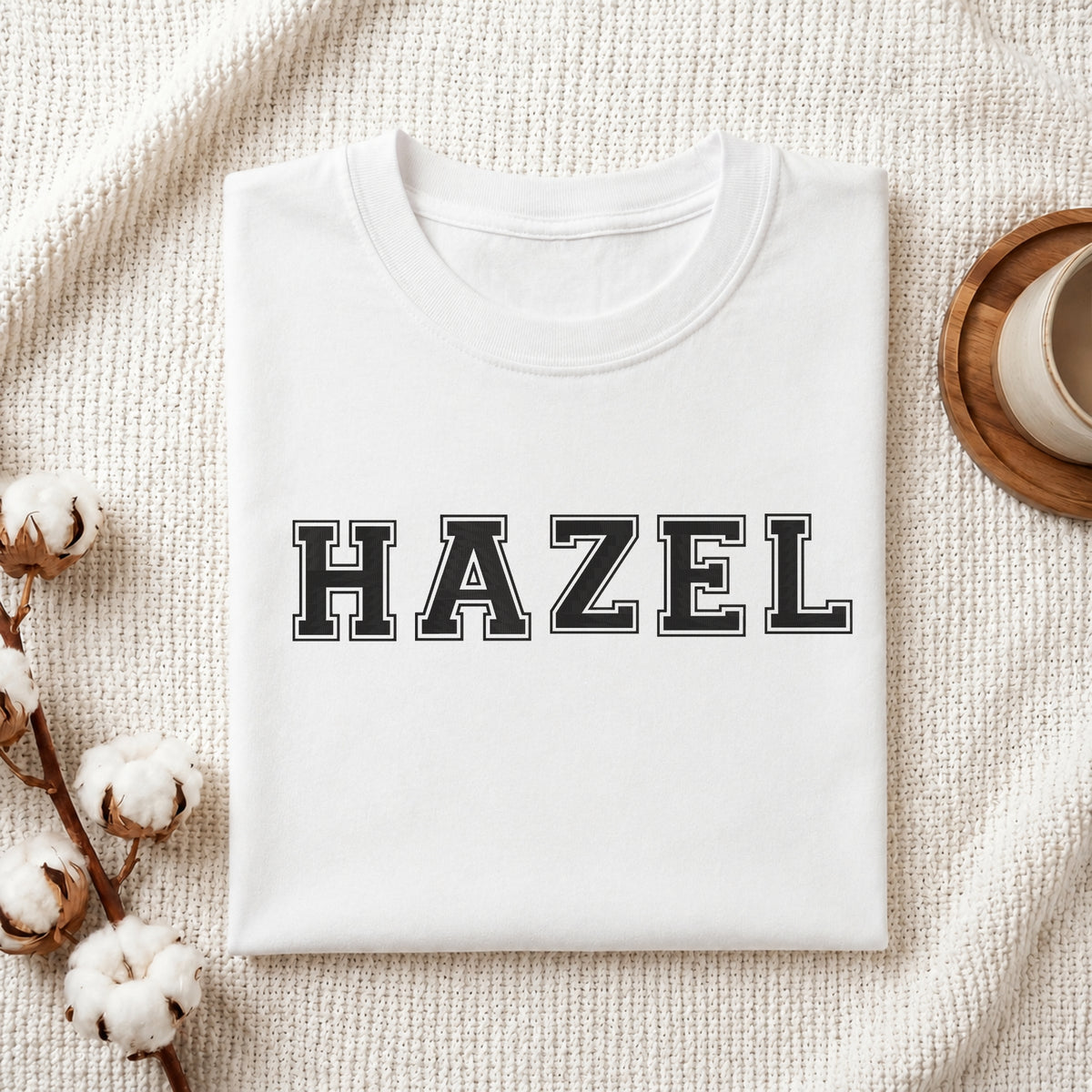 Custom name "HAZEL" stitched in varsity style black letters with white outline on a white folded cotton t-shirt with cotton branch decoration.