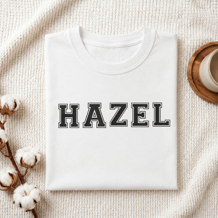Custom name "HAZEL" stitched in varsity style black letters with white outline on a white folded cotton t-shirt with cotton branch decoration.