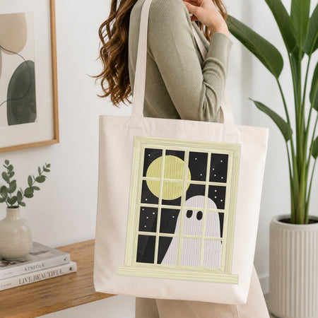 A woman wearing a sage green ribbed sweater carries a cream canvas tote bag with a ghost window graphic. The graphic features a pale yellow full moon, a white ghost, and a black starry night sky.