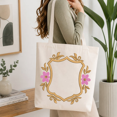 A woman wearing a sage green ribbed sweater carries a cream canvas tote bag with a gold ornate frame and pink flowers on the front, set in a bright, modern room with a wooden table and potted plants.