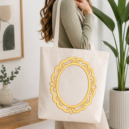 A woman wearing a sage green ribbed sweater carries a cream canvas tote bag with a large yellow ornate frame graphic on the front, set in a bright, modern room with a wooden table, books, and a potted plant.