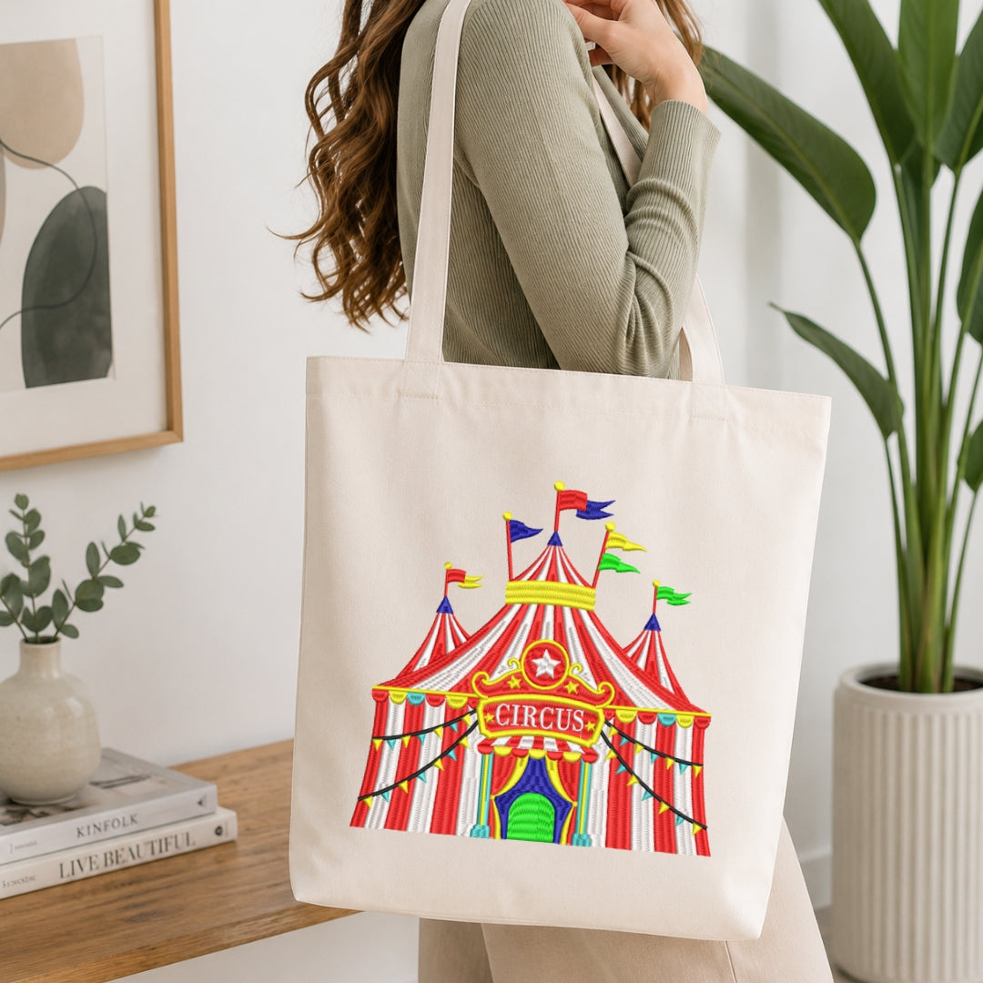 A woman wearing a sage green ribbed long-sleeve top carries a cream canvas tote bag featuring a colorful circus tent graphic. The setting is a bright, modern room with a wooden side table, a framed print, a small potted plant, and a larger green houseplant in the background.