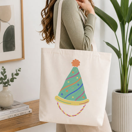 A woman wearing a sage green ribbed long-sleeve top carries a cream canvas tote bag with a colorful party hat illustration. The background shows a bright, modern room with a wooden table, books, a framed print, and a potted plant.