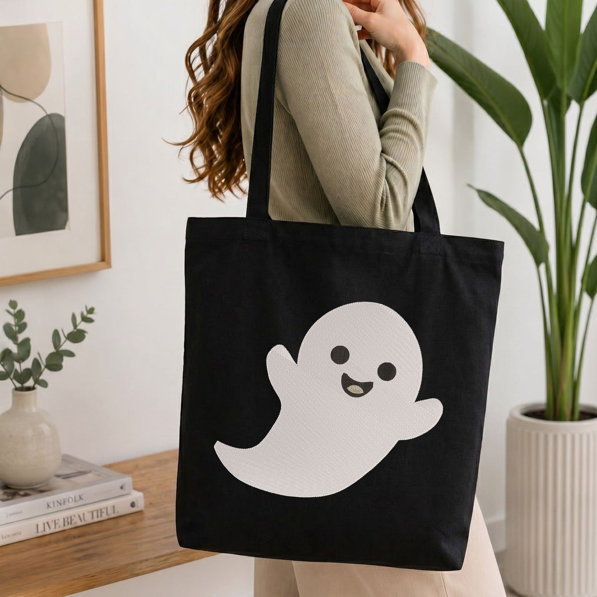 A person wearing a beige ribbed sweater carries a black canvas tote bag with a large white cartoon ghost graphic on the front, set in a bright indoor room with a plant and framed art in the background.