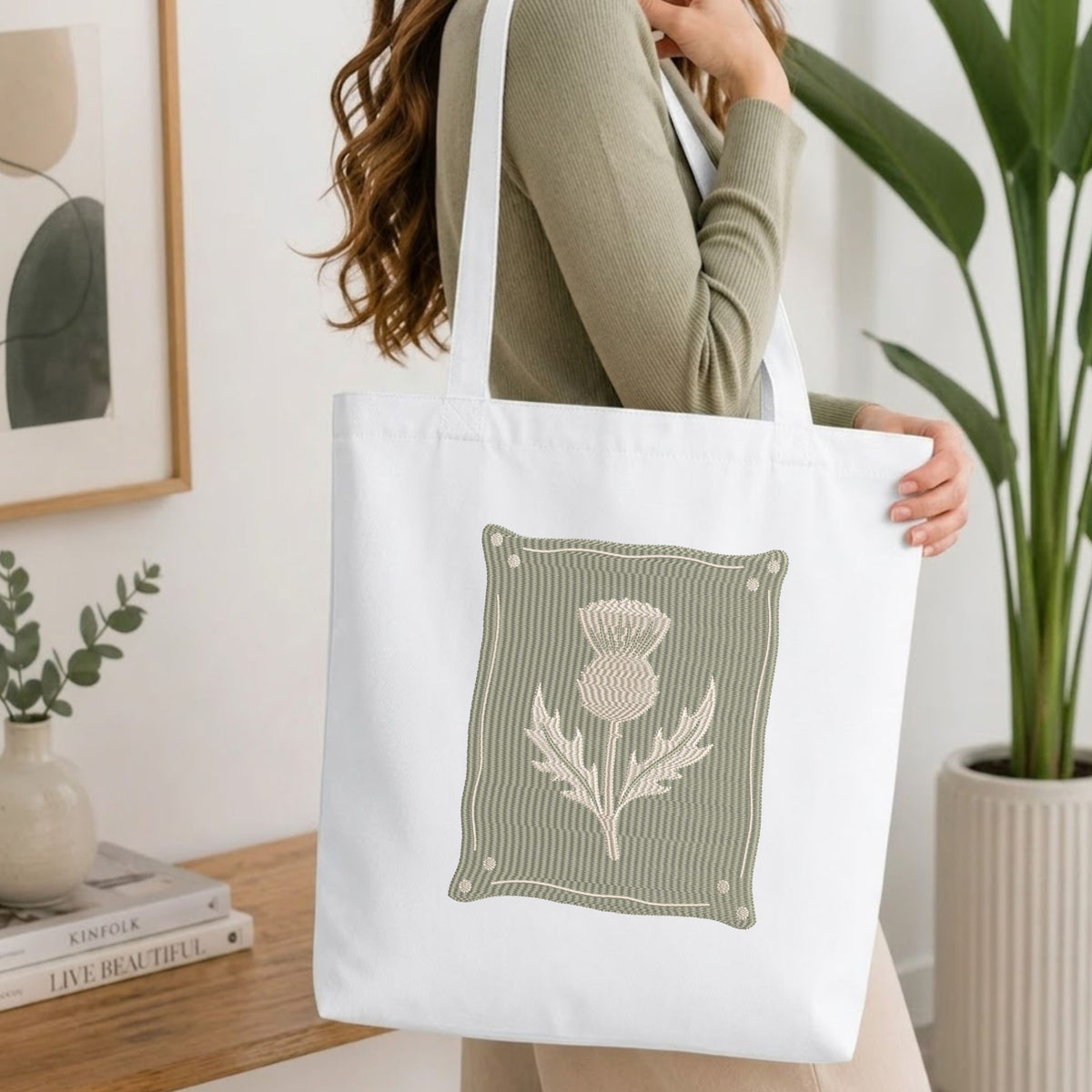 A woman wearing a sage green ribbed long-sleeve top carries a white canvas tote bag with a large embroidered thistle design on the front. The setting is a bright, modern room with a wooden table, books, a framed print, and a potted plant.