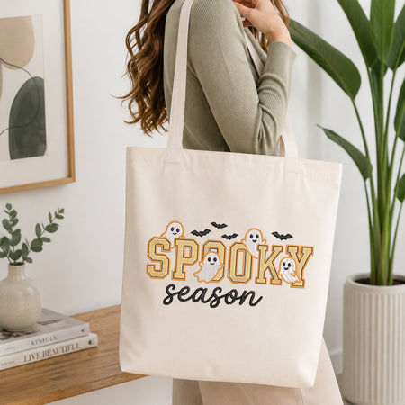 A woman wearing a sage green ribbed long-sleeve top carries a cream canvas tote bag with a 'Spooky Season' Halloween graphic. The graphic features the word 'Spooky' in a textured gold font with white ghosts and black bats, and 'season' in black script.