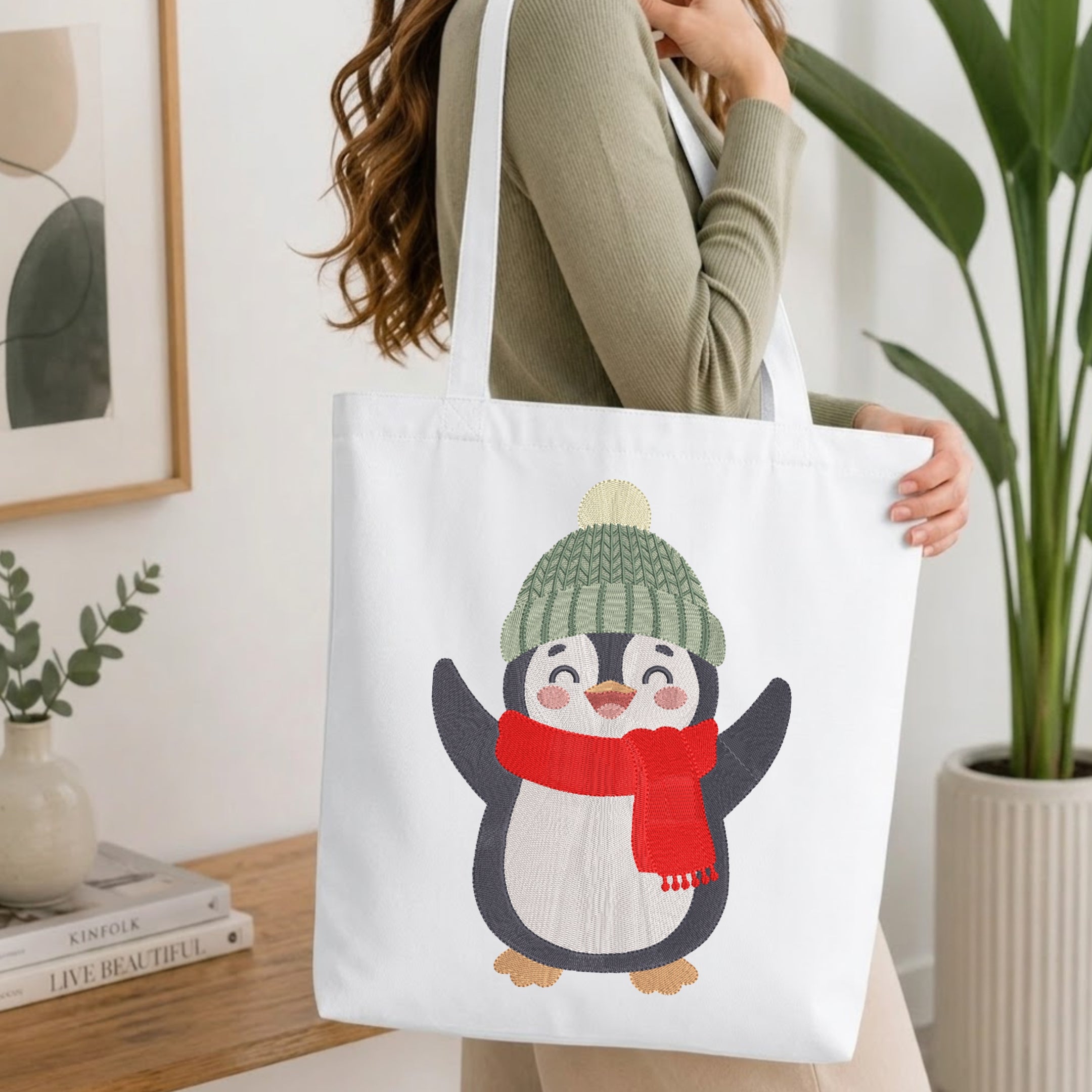 A woman wearing a sage green ribbed sweater carries a white canvas tote bag featuring a cute cartoon penguin wearing a green knit hat and red scarf, set against a bright, modern indoor background with a wooden table and potted plants.