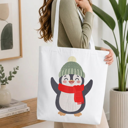 A woman wearing a sage green ribbed sweater carries a white canvas tote bag featuring a cute cartoon penguin wearing a green knit hat and red scarf, set against a bright, modern indoor background with a wooden table and potted plants.
