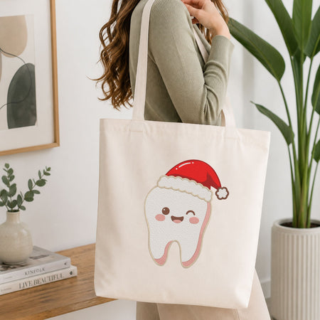 Woman carrying a white natural canvas tote bag featuring a cute winking tooth wearing a Santa hat Christmas dental machine embroidery design in a bright, modern interior setting  