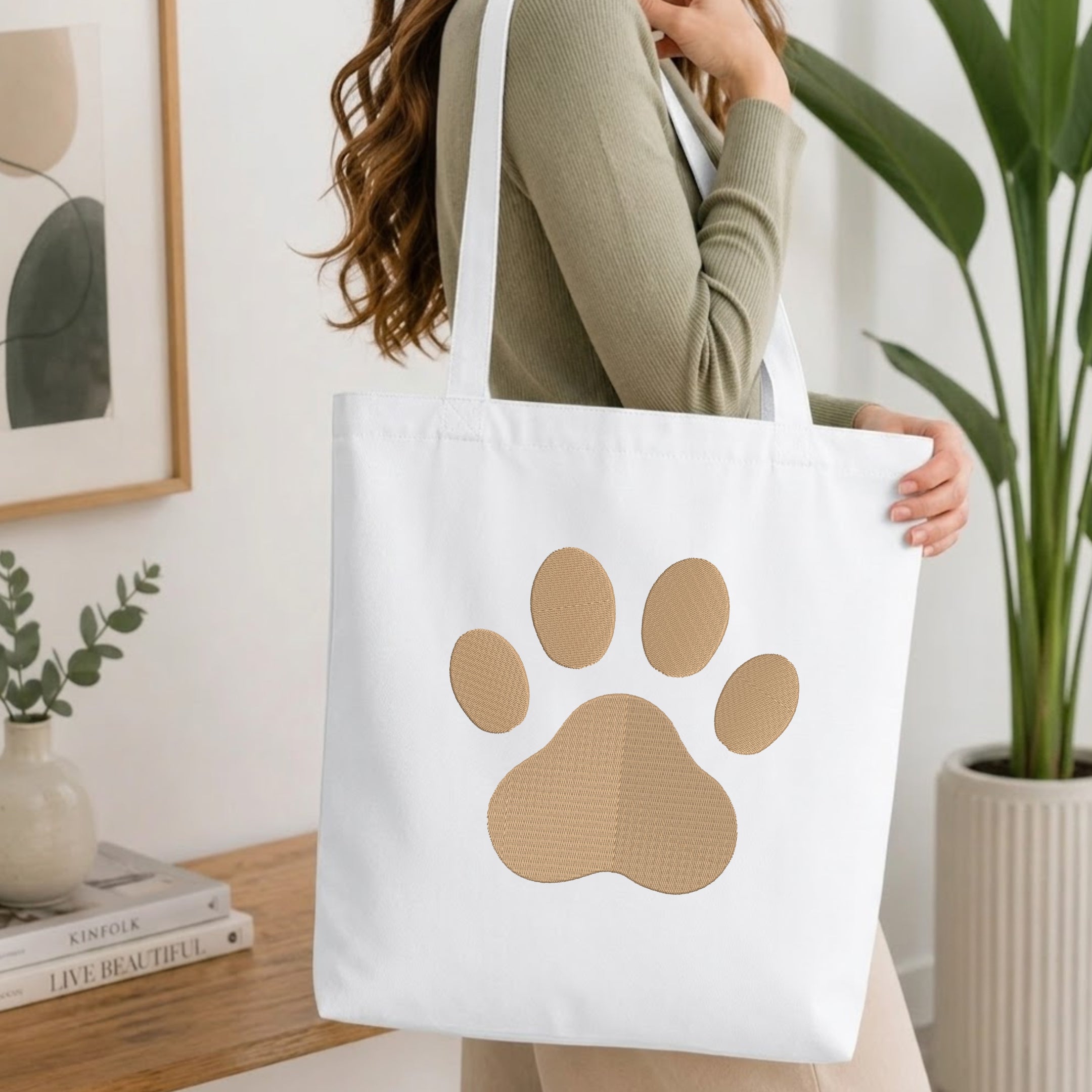 A woman wearing a sage green ribbed sweater carries a white canvas tote bag with a large tan paw print graphic on the front, set in a bright, modern room with a wooden table and potted plants.